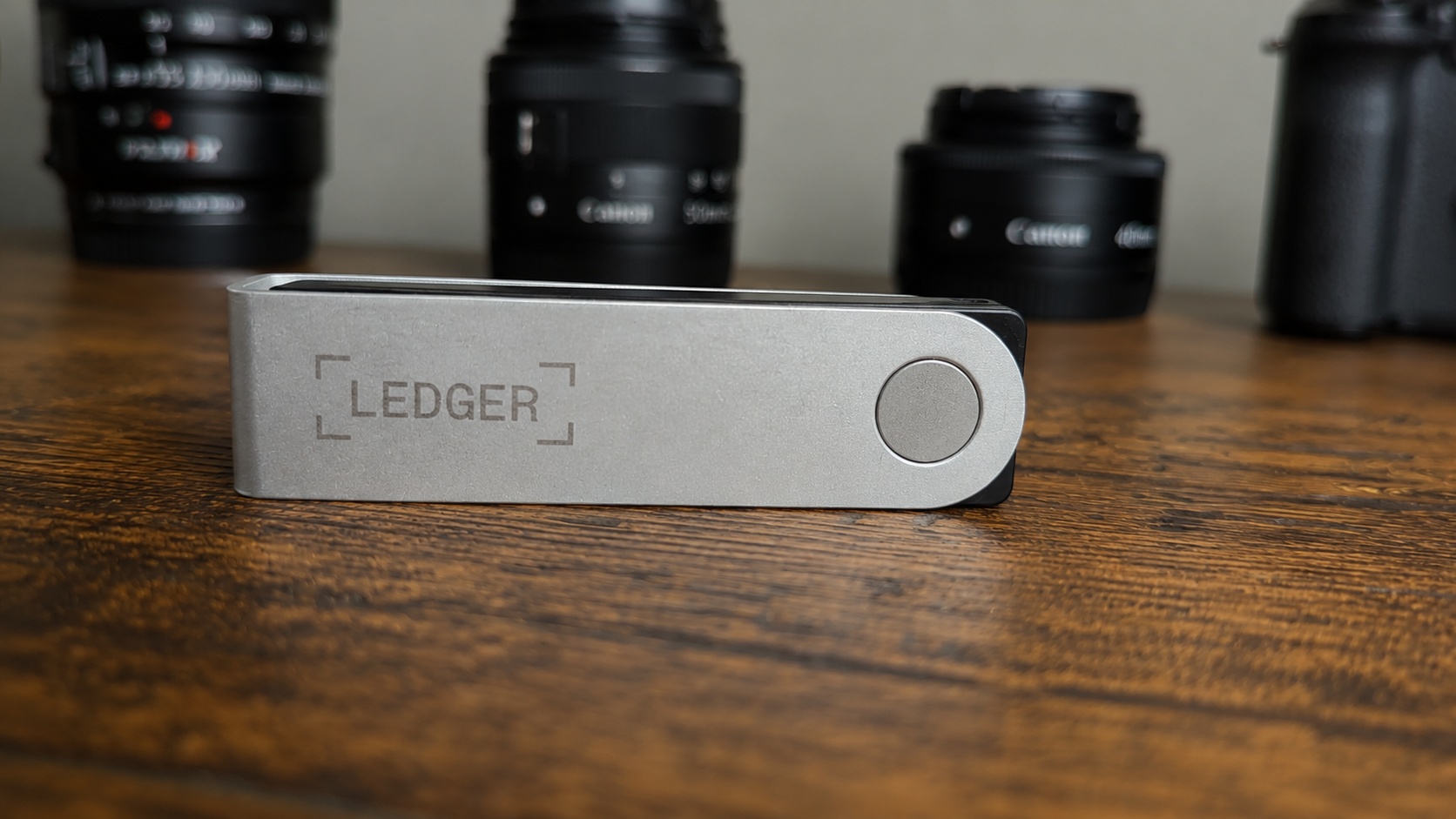 Ledger Nano X hardverski kripto novčanik na drvenom stolu sa objektivima u pozadini
